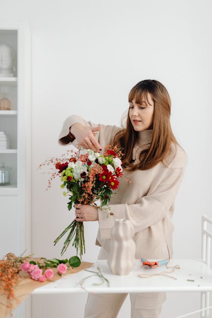 A young woman with long, light brown hair and bangs is arranging a vibrant floral bouquet indoors. She is wearing a beige, oversized sweater and appears focused as she adjusts the bouquet with her rig