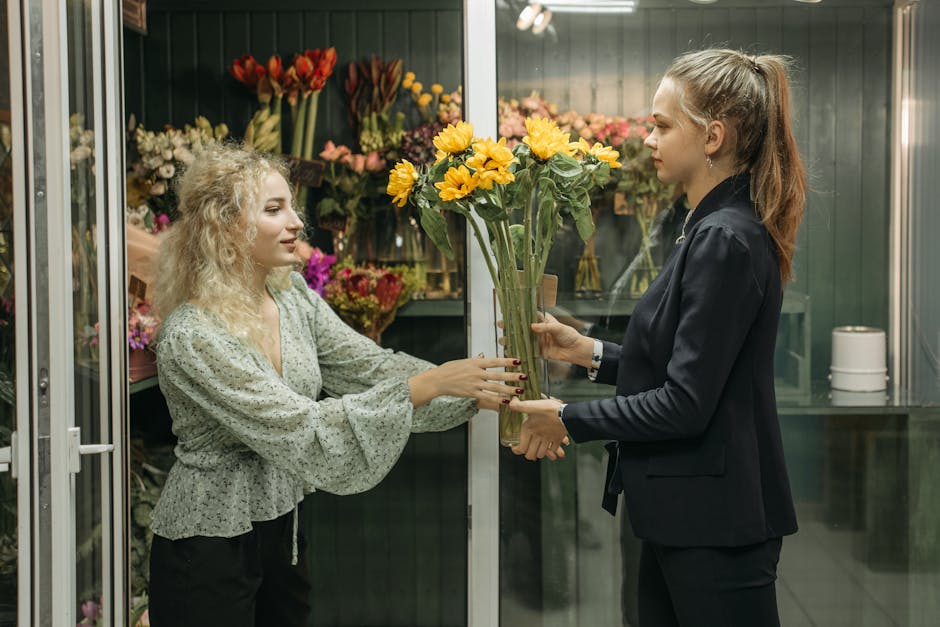 A florist with blonde, curly hair and a light green floral blouse is handing over a bouquet of bright yellow sunflowers to a customer, a young woman with long brown hair tied in a high ponytail, dress