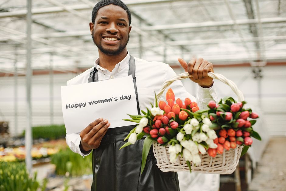 A smiling male florist in a greenhouse holds a wicker basket filled with fresh tulips in shades of pink, white, and coral, arranged with green foliage. He is wearing a white shirt and a black apron, a