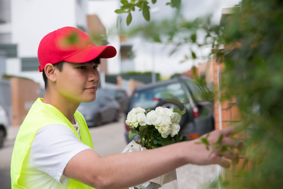 A young male florist delivering a floral gift with a bouquet of white hydrangeas wrapped in white paper, holding the arrangement close to his chest as he prepares to hand it over. He is wearing a brig